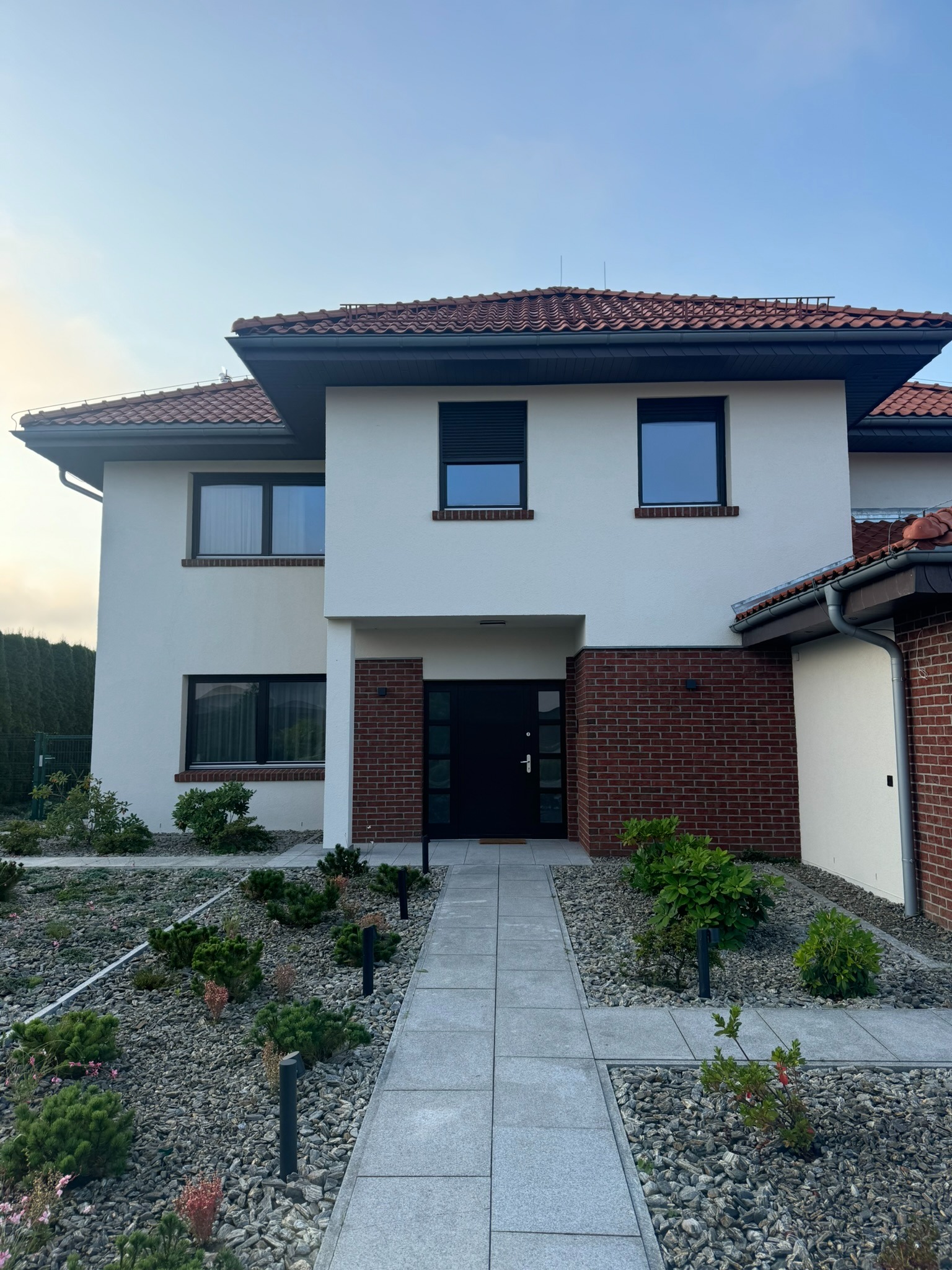 Biała elewacja domu z ceglanymi akcentami, czarnymi oknami i drzwiami. Uwagę przykuwa zadbany ogród z kamieniami i niskimi krzewami oraz oświetlenie ścieżki.