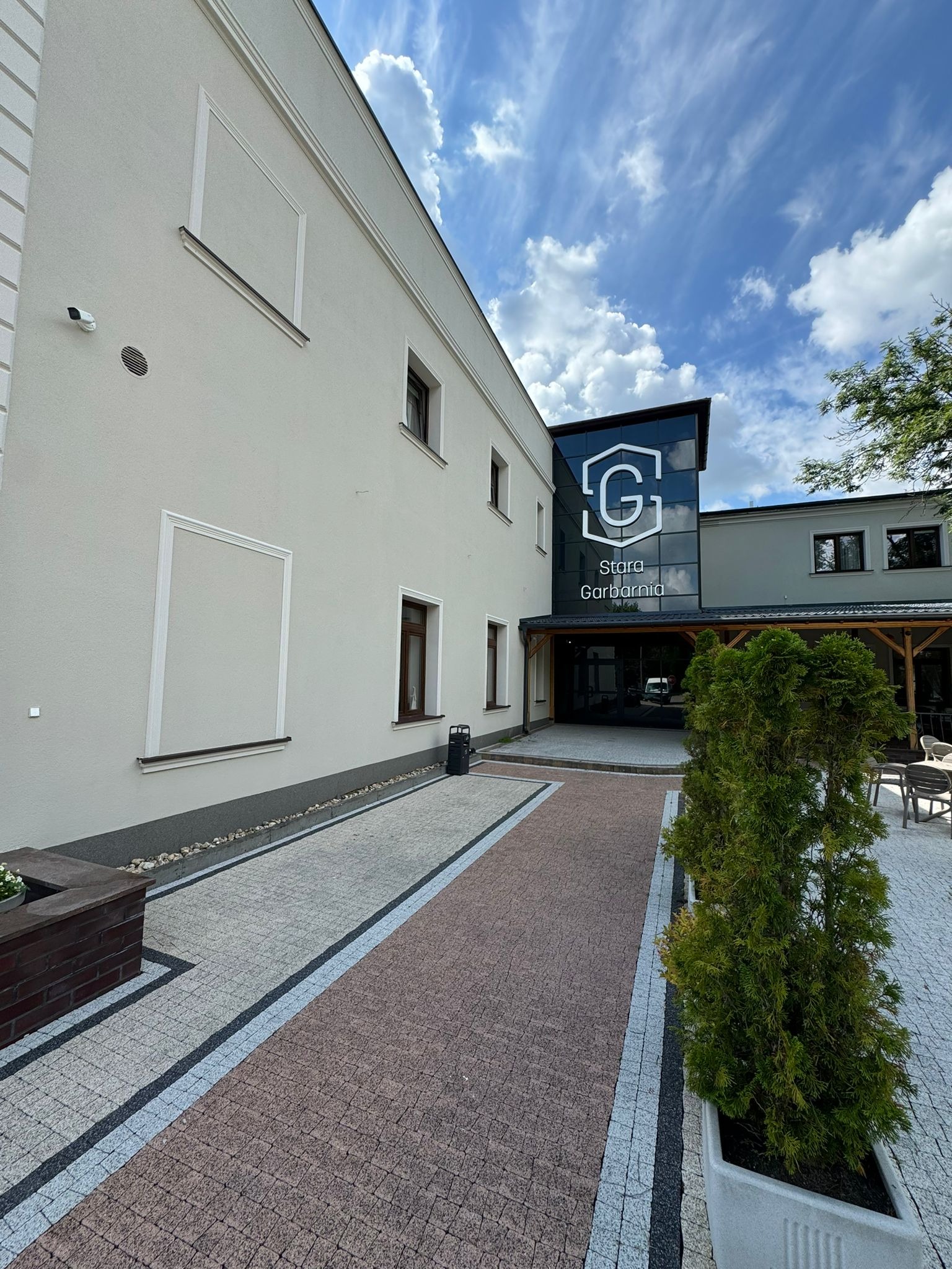 Jasna elewacja budynku 'Stara Garbarnia' z nowoczesnym, przeszklonym wejściem. Uwagę przyciąga świeżo pomalowana fasada i zadbany chodnik z kostki.