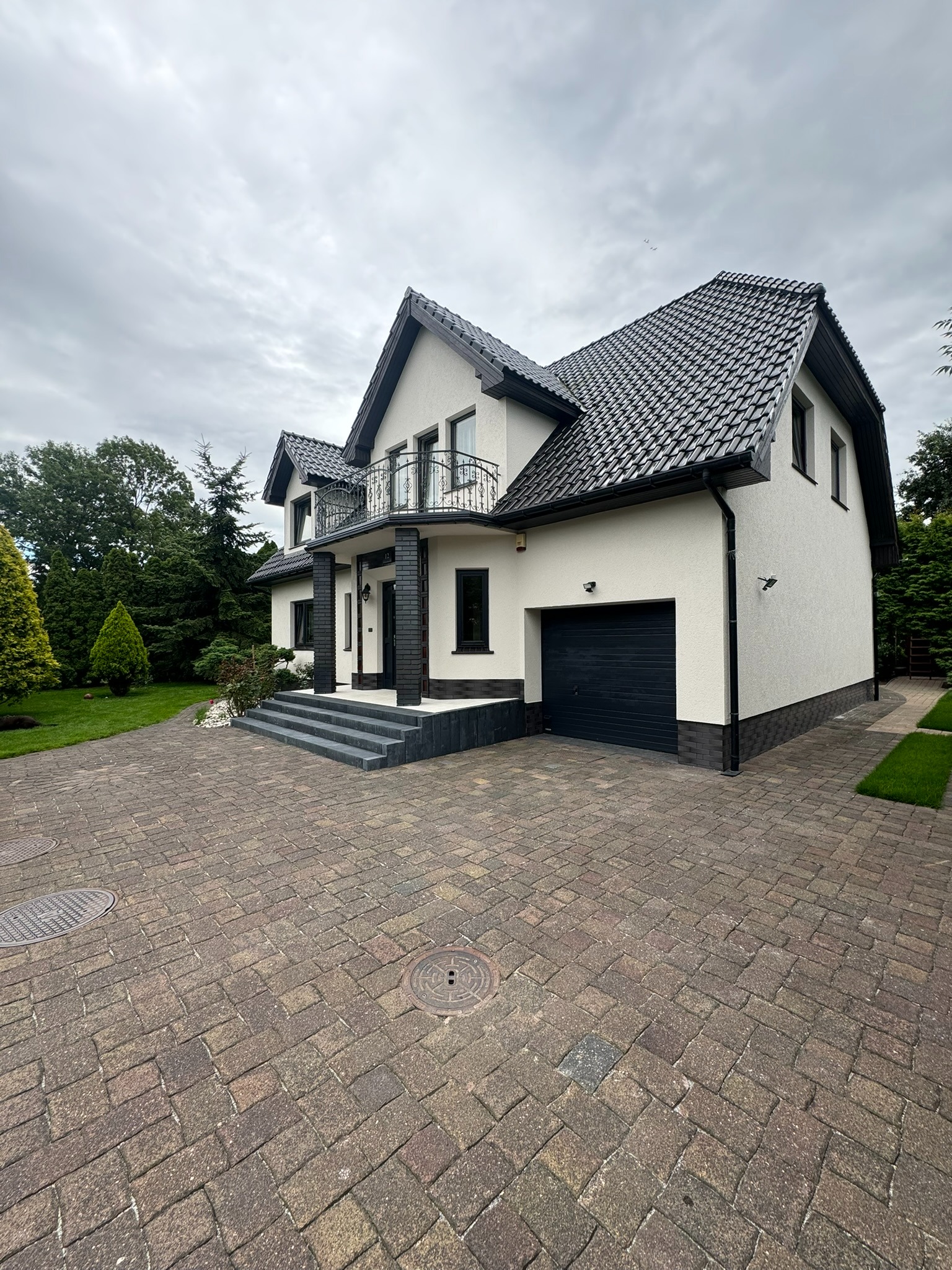 Elegancki dom z białą elewacją i czarnym dachem, widok z brukowanego podjazdu. Balustrada balkonu z kutego żelaza. Minimalistyczny design z akcentami zieleni.