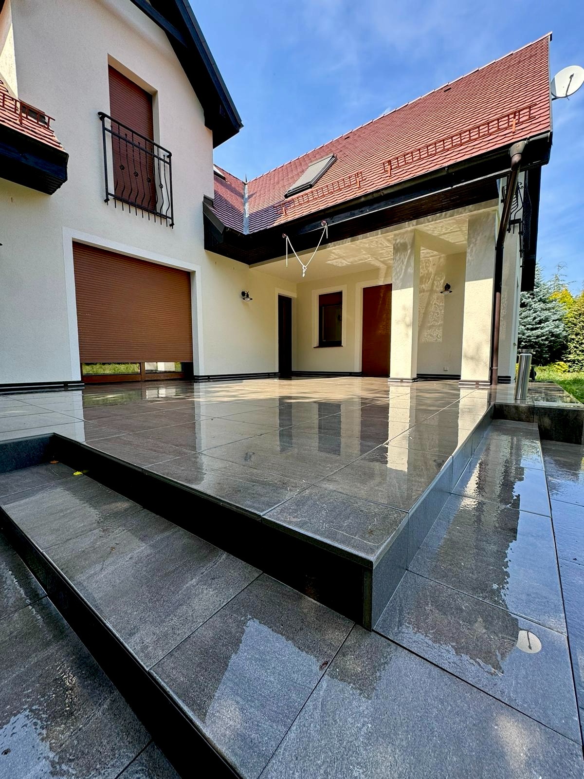 Elegancki dom z jasną elewacją i nową, błyszczącą, szarą terakotą na tarasie. Widoczne schody wejściowe i fragment ogrodu. Czyste, odbijające światło płytki.