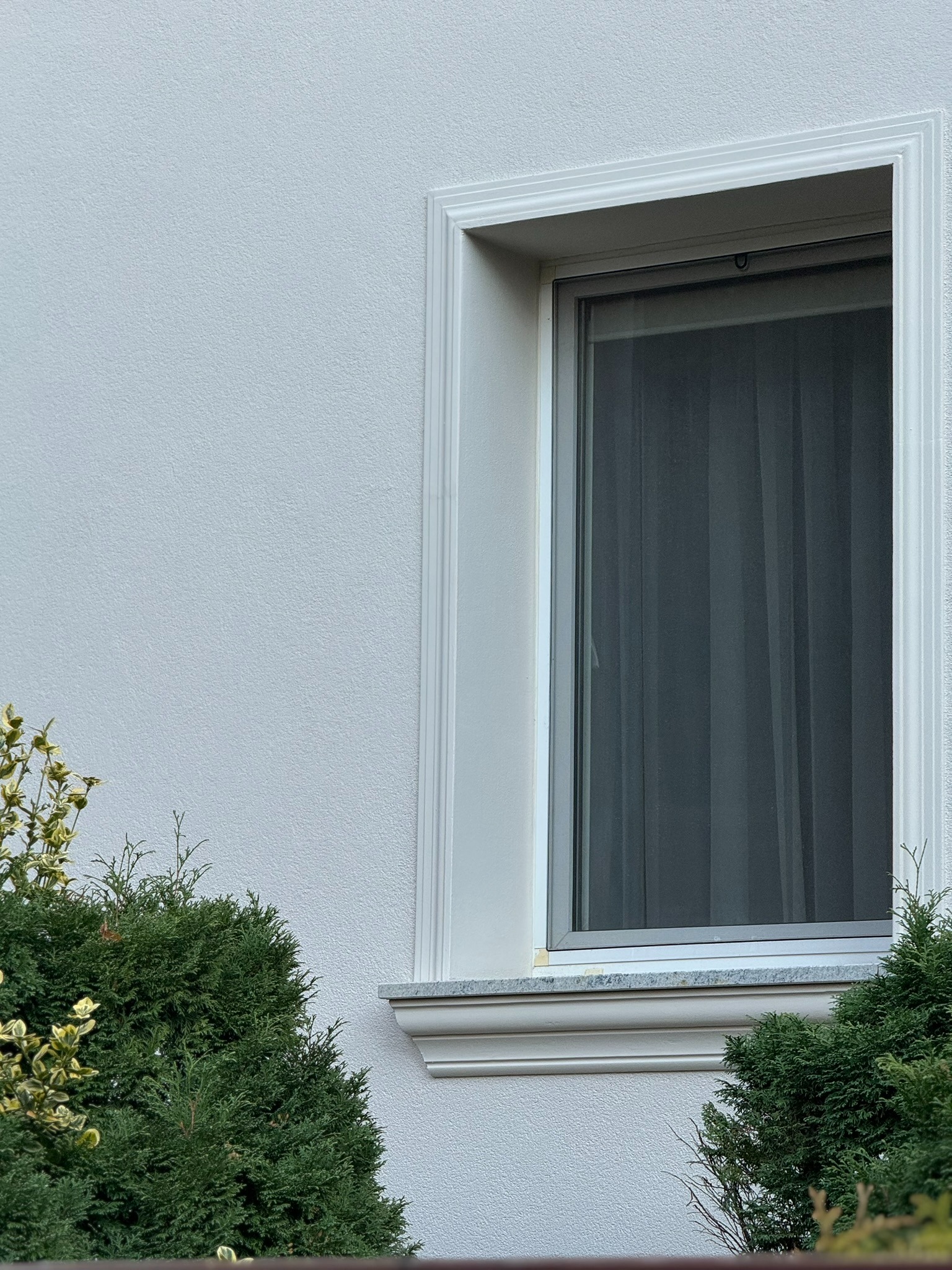 Biała elewacja z oknem w ozdobnej ramie, widok z dołu, krzewy w tle. Detal wykończenia budynku we Wrocławiu.