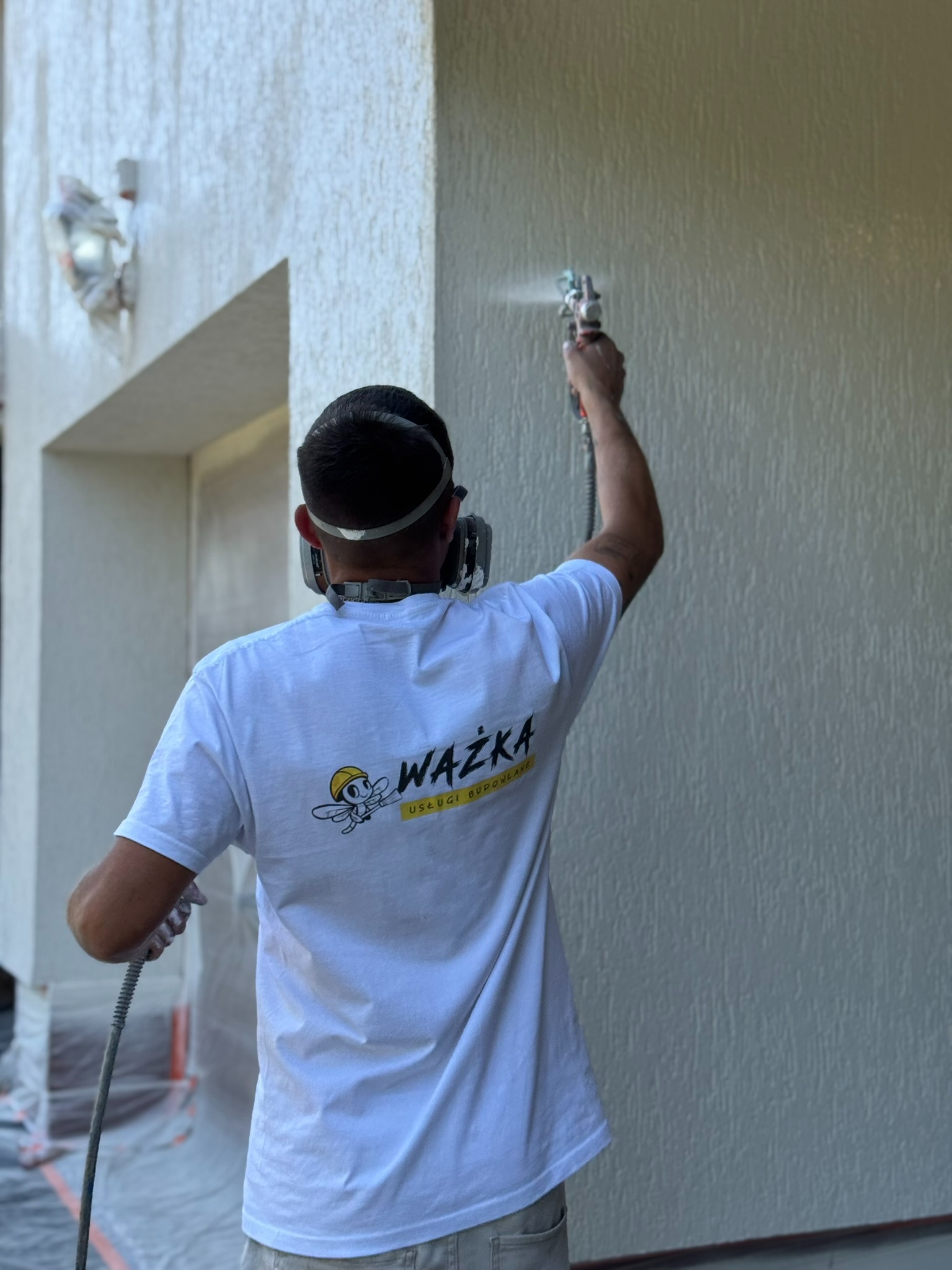 Malowanie elewacji natryskowo przez pracownika w masce i koszulce z logo firmy. Widoczna struktura elewacji i zabezpieczone elementy.