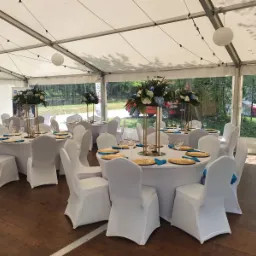 Uroczyste stoły z białymi obrusami i krzesłami w pokrowcach, złote talerze i turkusowe serwetki, wysokie kompozycje kwiatowe w złotych wazonach, widok na zewnątrz z zalesionym tłem przez...