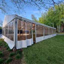 Duży, biały namiot z przezroczystymi ścianami, oświetlony girlandami żarówek, ustawiony na zielonej trawie w Rzeszowie, widoczne drzewa w tle i stoliki z parasolami po lewej stronie.