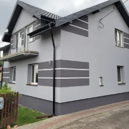 Dwukondygnacyjny dom z szarą elewacją, ozdobiony poziomymi, białymi pasami, z czarną, metalową rynną i dachem z blachy trapezowej. Na pierwszym planie brązowy płot z numerem domu 12.