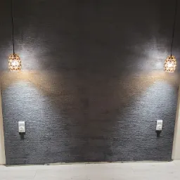 Ciemnoszara ściana z wyraźną fakturą, oświetlona dwoma wiszącymi lampami o ażurowych kloszach, z widocznymi gniazdkami elektrycznymi po bokach, na tle białych ścian i jasnej podłogi.