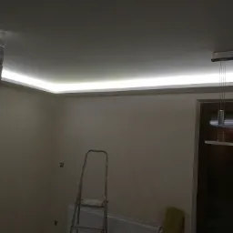 Wnętrze pokoju z podwieszanym sufitem, oświetleniem LED wzdłuż krawędzi, dekoracyjną ścianą z kamienia, drabina oparta o ścianę, grzejnik i lampa wisząca.