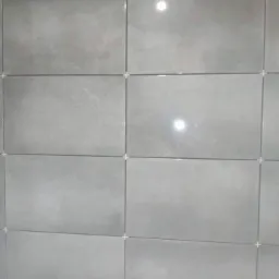 Ściana wyłożona szarymi, prostokątnymi płytkami ceramicznymi z krzyżykami dystansowymi w spoinach, widoczne odbicia światła.