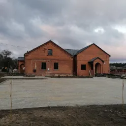 Odrestaurowany budynek z czerwonej cegły z nową kostką brukową na podwórzu, widoczne młode drzewka, pochmurne niebo.