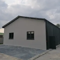 Nowo wybudowany budynek o szarej elewacji z czarnymi oknami i bramą, wyłożony kostką brukową, z widocznym fragmentem samochodu i pochmurnym niebem.