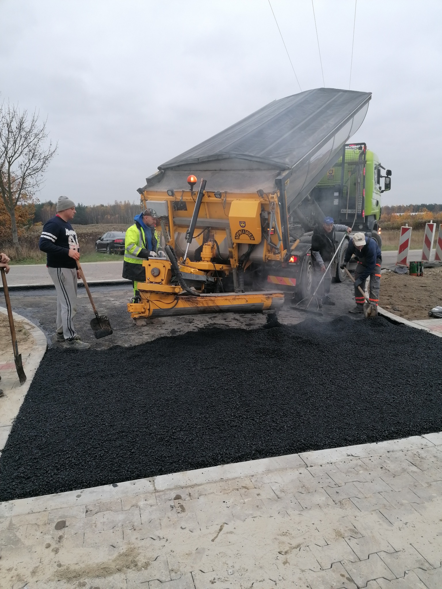 Układanie asfaltu na drodze przy użyciu rozściełacza i pracy ręcznej, widoczny zielony samochód ciężarowy w tle, pochmurne niebo.