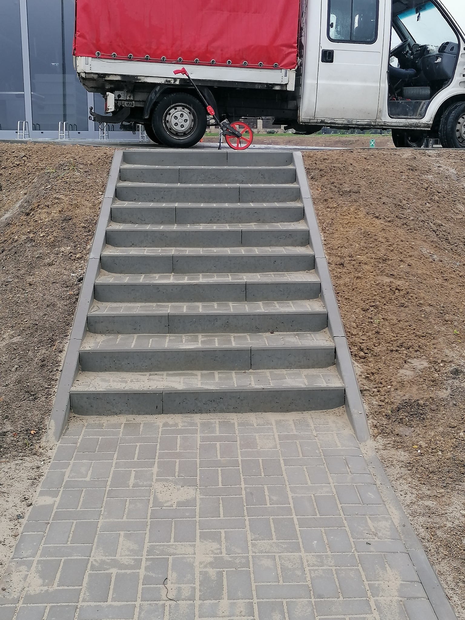 Nowe, betonowe schody prowadzące z chodnika z kostki brukowej do podwyższonego terenu, z zaparkowanym białym samochodem dostawczym z czerwoną plandeką w tle.