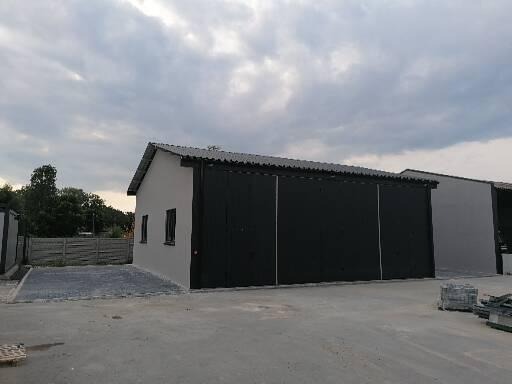 Nowo wybudowany, parterowy budynek gospodarczy z czarną, przesuwną bramą i jasnoszarą elewacją, widoczny na tle pochmurnego nieba.