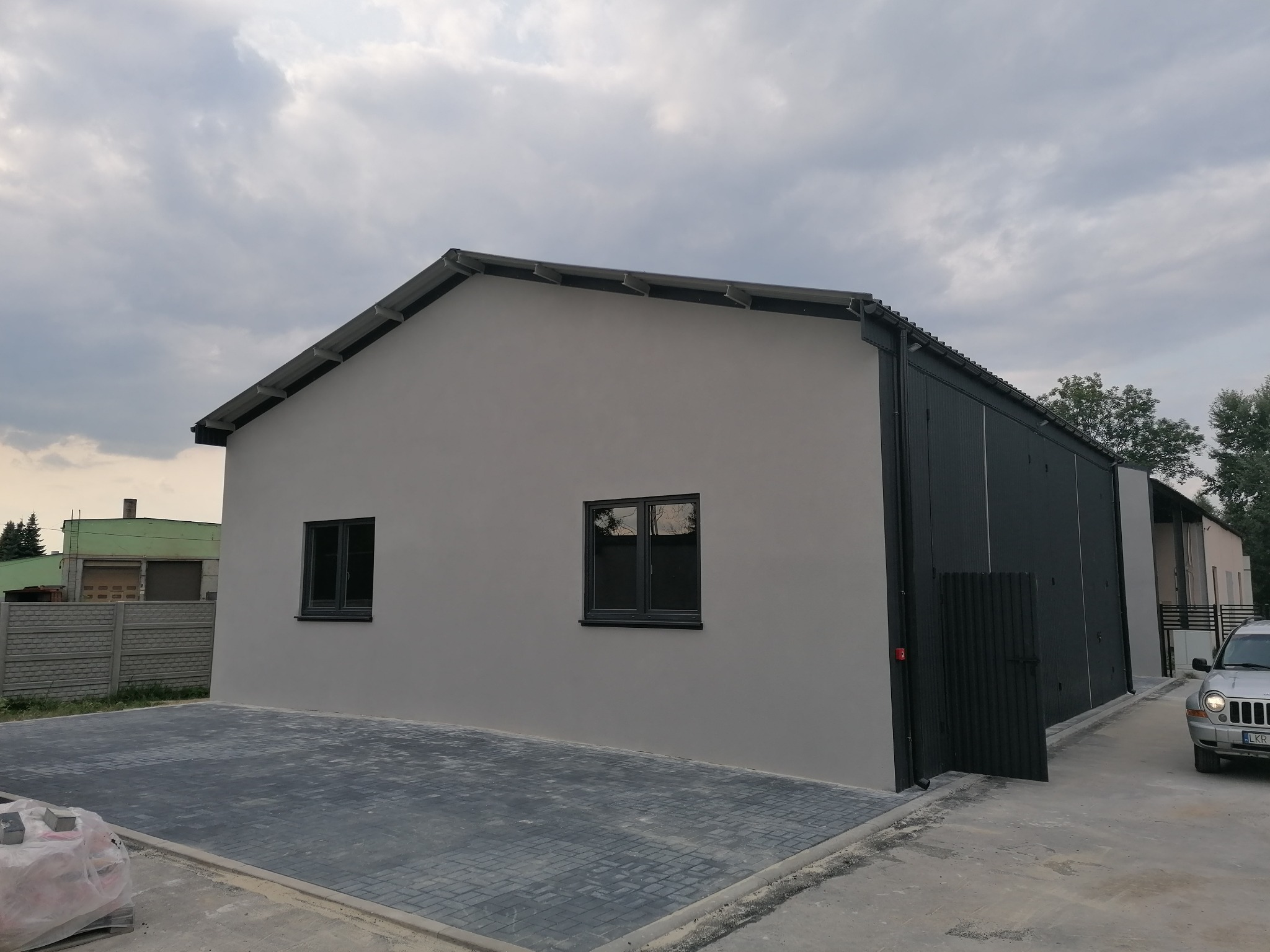 Nowo wybudowany budynek o szarej elewacji z czarnymi oknami i bramą, wyłożony kostką brukową, z widocznym fragmentem samochodu i pochmurnym niebem.