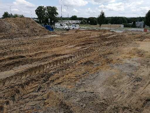 Przygotowany teren pod budowę z widocznymi śladami opon maszyn budowlanych, w tle zaparkowane samochody dostawcze i drzewa.