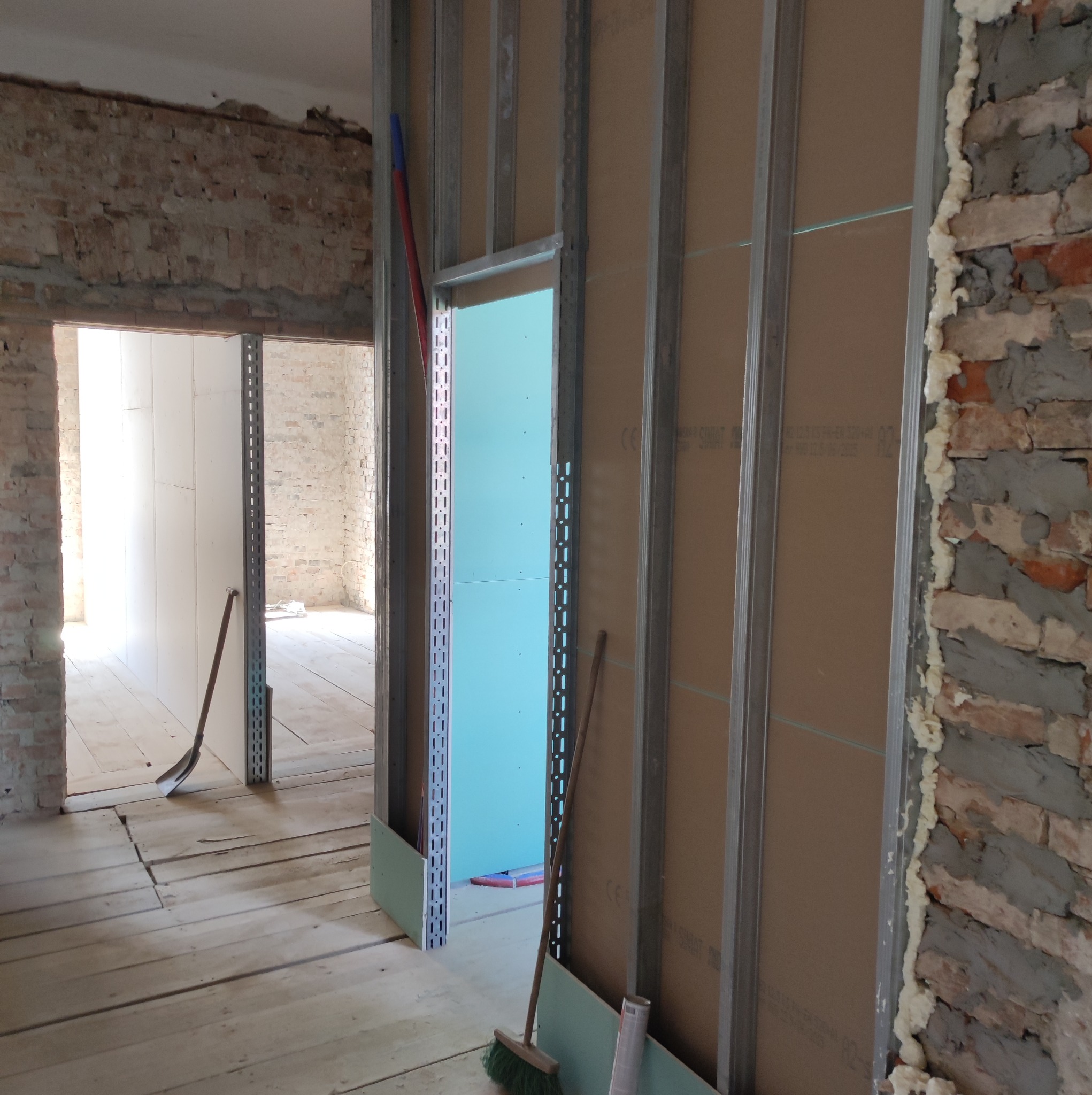 Wnętrze w trakcie prac wykończeniowych: metalowy szkielet ścianki działowej z płytami gipsowo-kartonowymi, fragment odsłoniętej ceglanej ściany z pianką montażową, drewniana podłoga, widoczne...