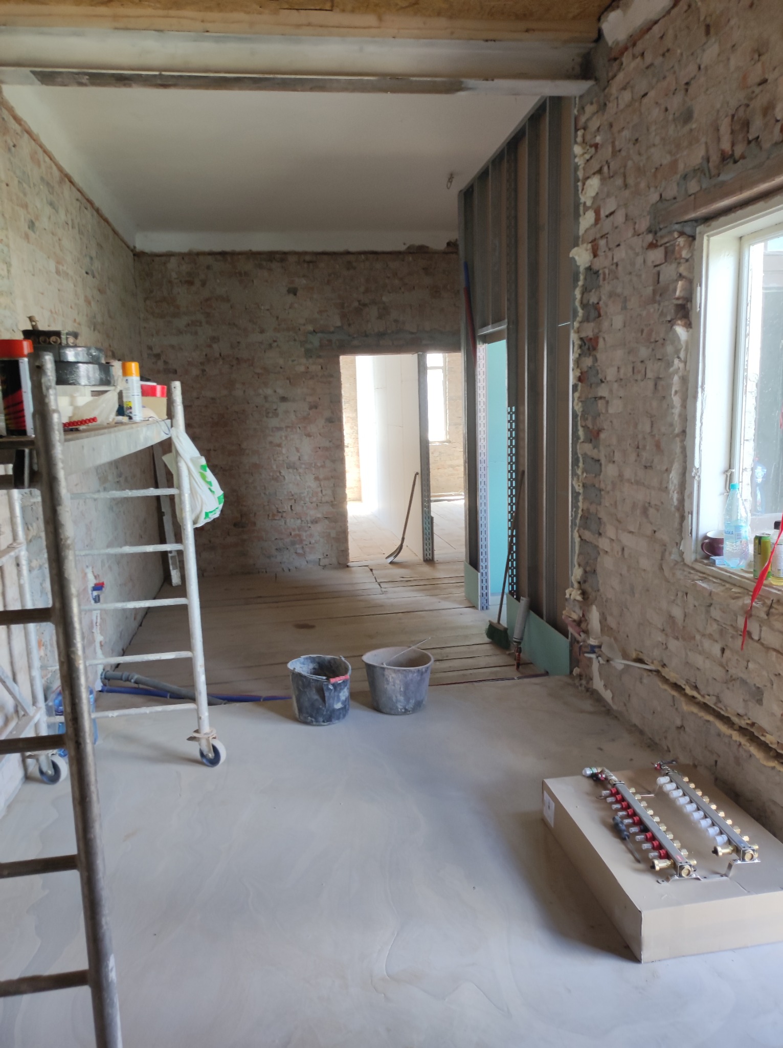 Wnętrze w trakcie remontu: surowe ceglane ściany, metalowa konstrukcja ścianki działowej, wylewka podłogowa, drabina malarska, wiadra z zaprawą i rozdzielacz ogrzewania podłogowego na kartonie.