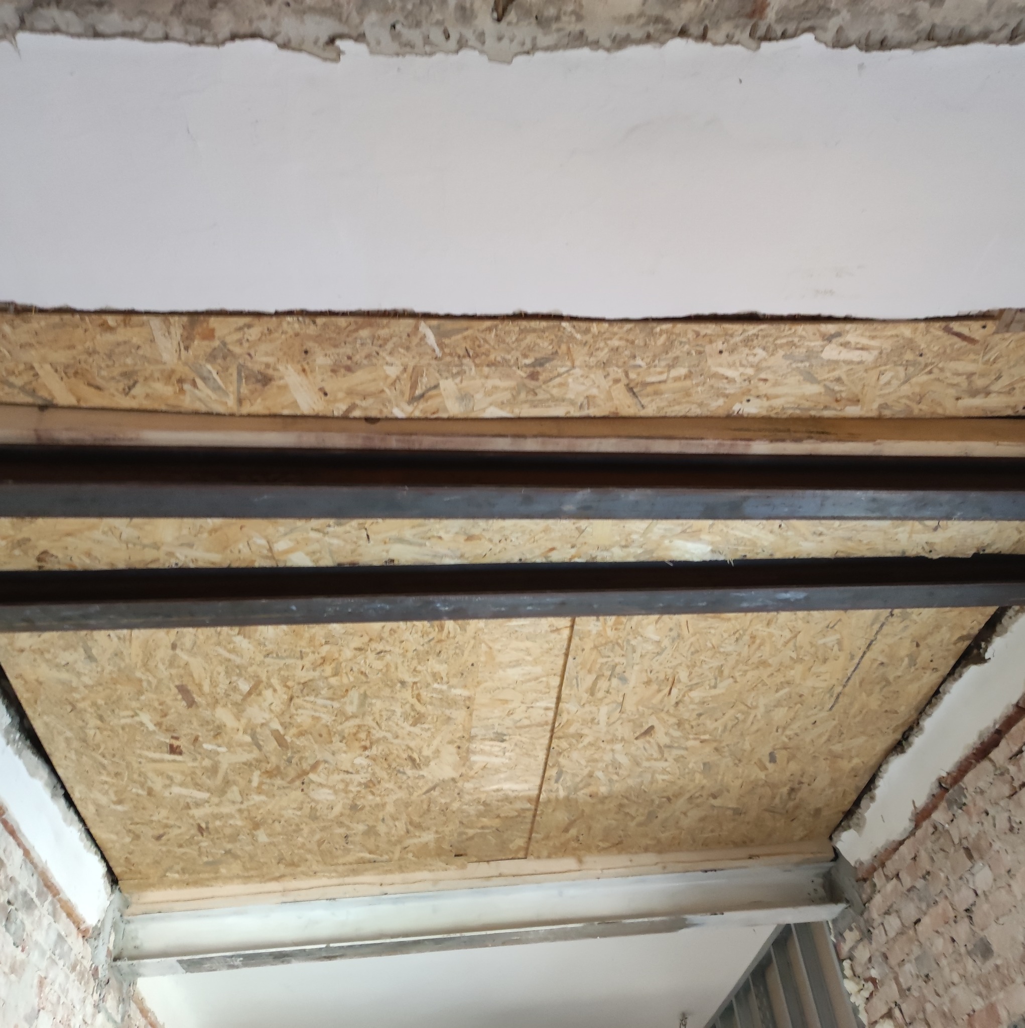 Montaż płyty OSB na metalowej konstrukcji pod sufitem, widoczne surowe wykończenie ściany i fragment instalacji.