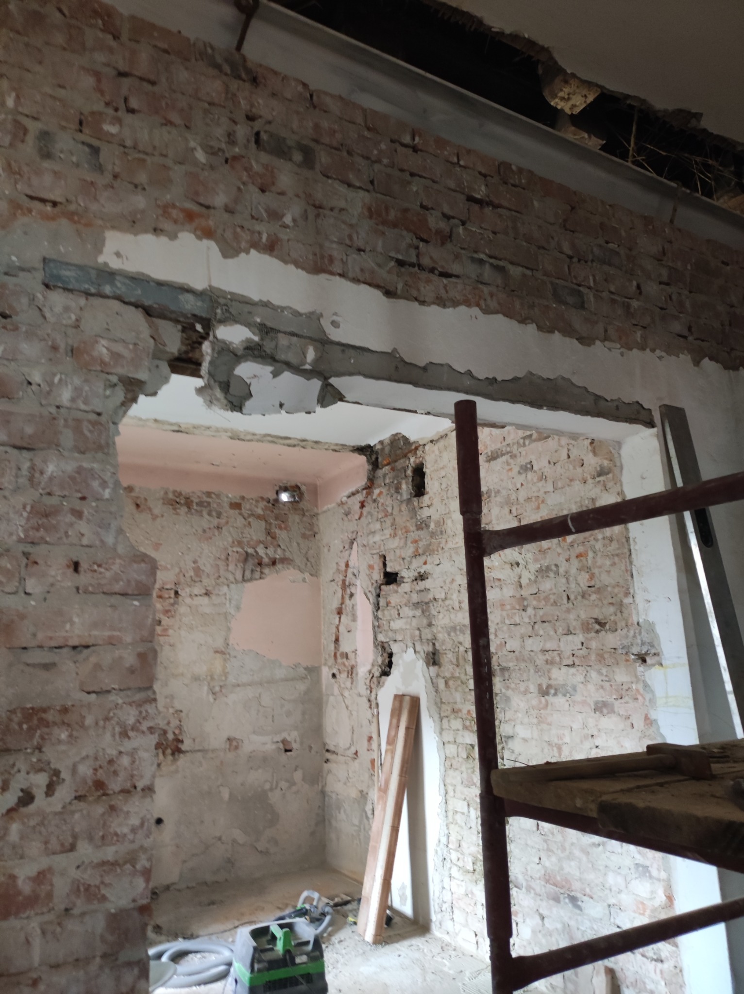 Wnętrze w trakcie demontażu: odsłonięta cegła, fragmenty tynku, stalowa belka nad otworem drzwiowym, rusztowanie i odkurzacz przemysłowy w tle