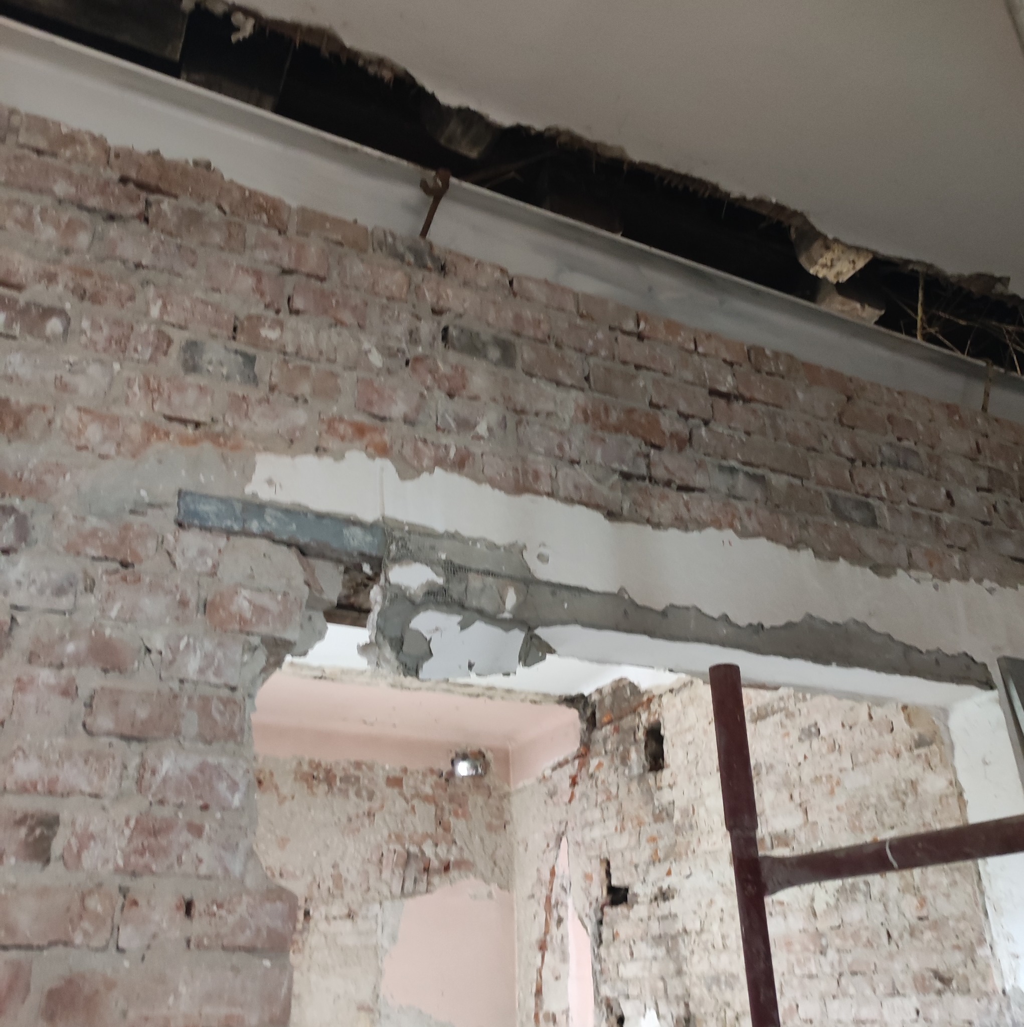 Ceglana ściana z widocznymi śladami po usuniętych elementach, odsłonięta metalowa belka pod sufitem i fragment rusztowania, ukazujące etap prac remontowych wewnątrz pomieszczenia.