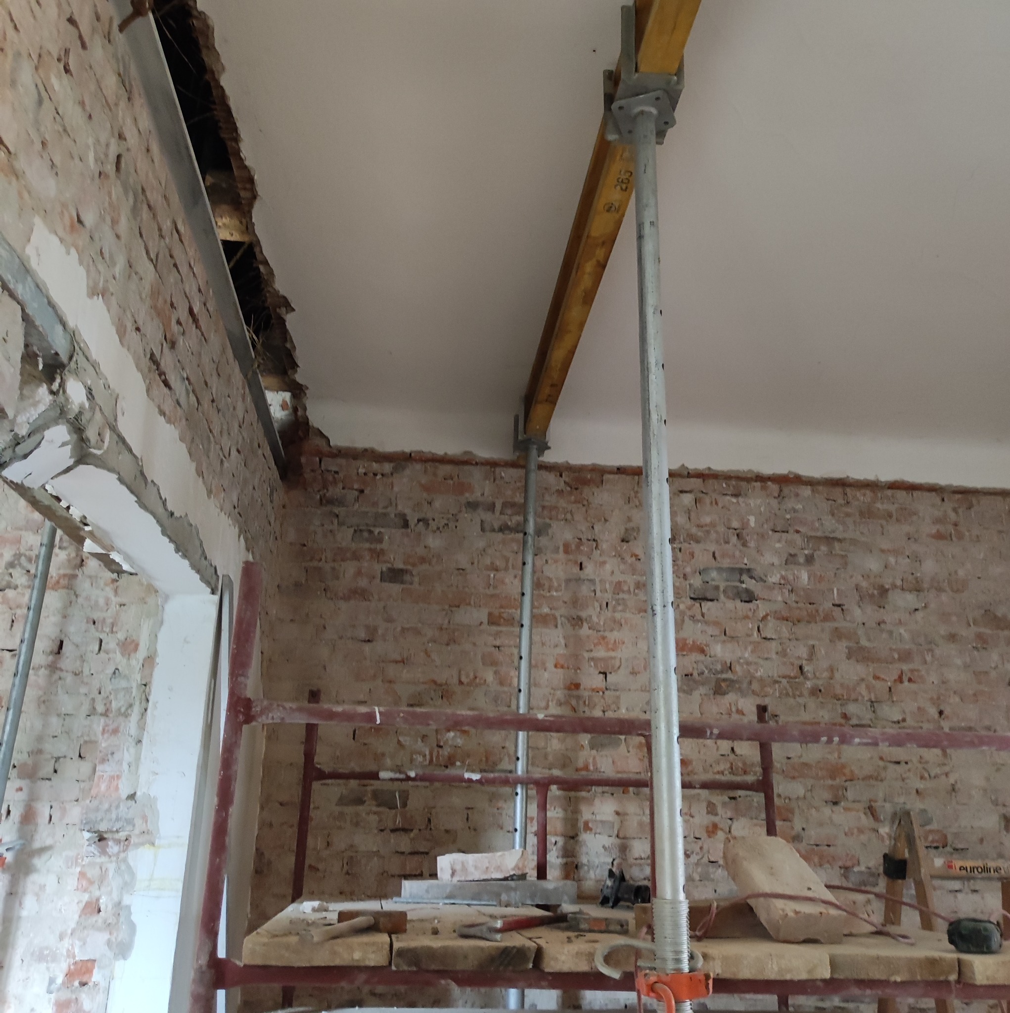 Wnętrze w trakcie remontu: odsłonięta ceglana ściana, podparte stropu metalowymi stemplami, widoczne rusztowanie i narzędzia na platformie.