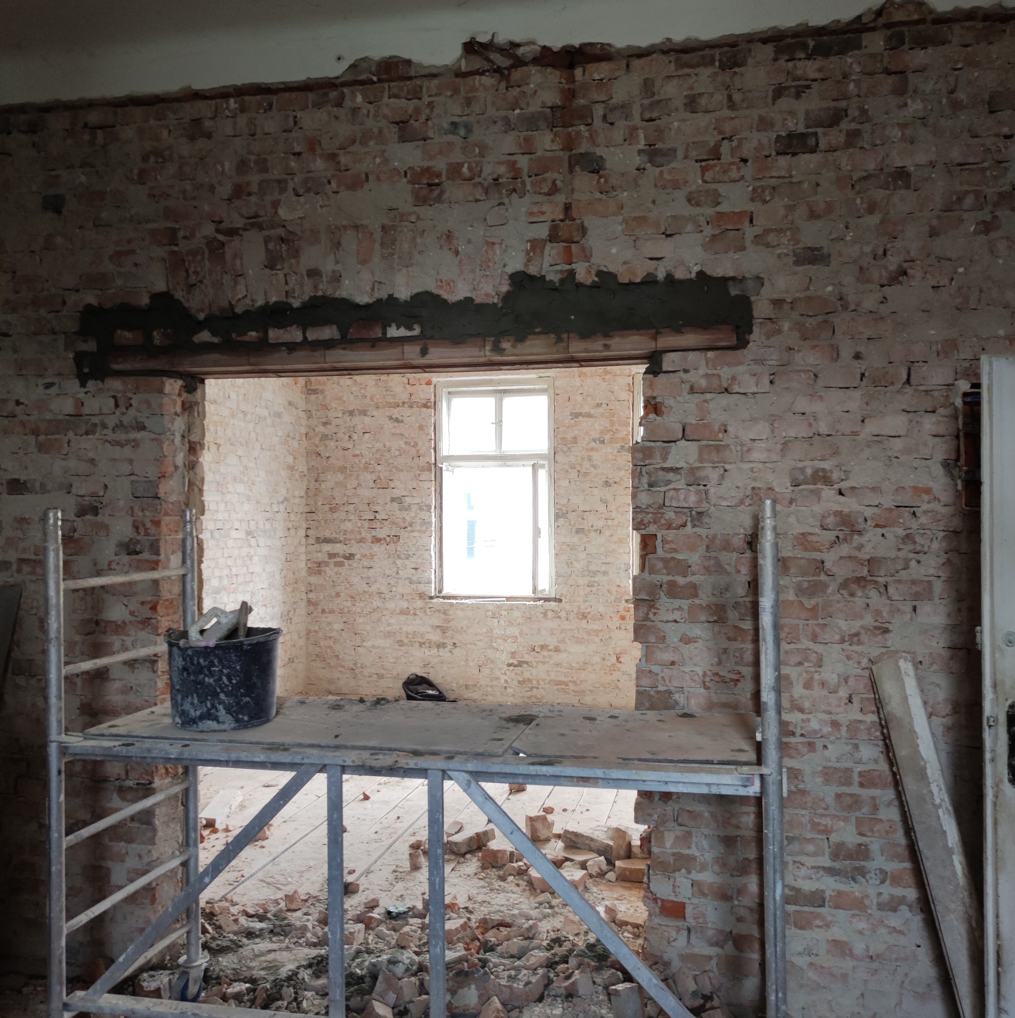 Ceglana ściana w trakcie demontażu z widocznym oknem w tle, rusztowanie z wiadrem i narzędziami na pierwszym planie, gruz na podłodze.