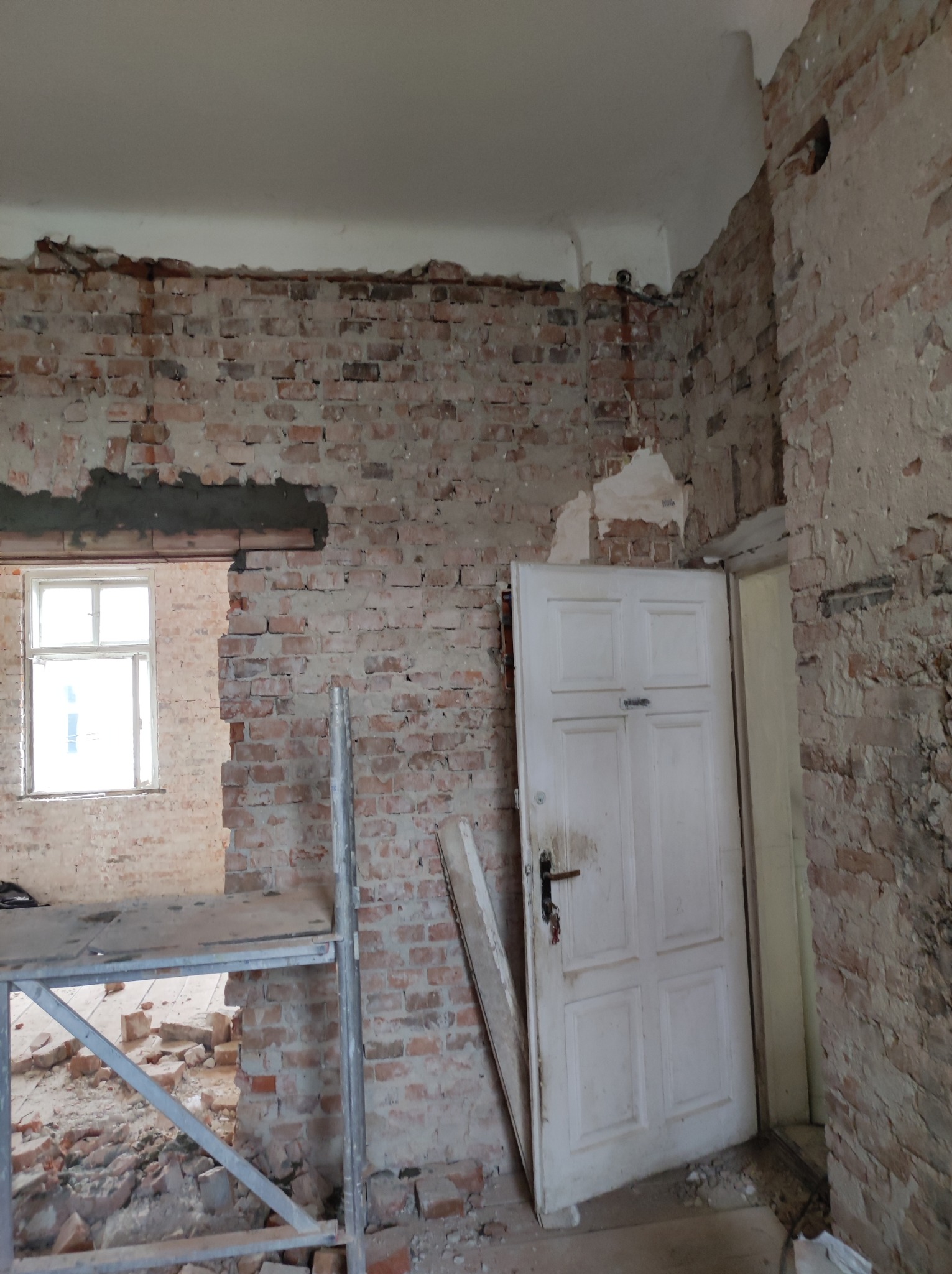 Wnętrze w trakcie demontażu: odsłonięta cegła, resztki tynku, białe drzwi wewnętrzne i fragment rusztowania. Widoczne pozostałości instalacji elektrycznej pod sufitem.