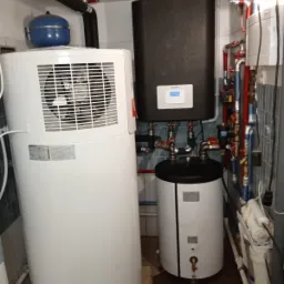 Kompaktowa kotłownia: biała pompa ciepła z wentylatorem, czarny zbiornik buforowy z wyświetlaczem, biały zasobnik ciepłej wody i liczne rury instalacji grzewczej w pomieszczeniu z szarymi płytkami.