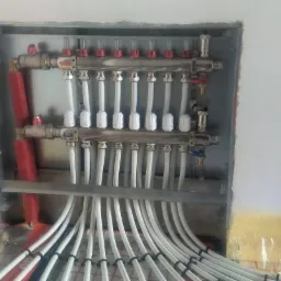 Rozdzielacz ogrzewania podłogowego z białymi rurami w metalowej obudowie w trakcie montażu, widoczna izolacja podłogowa z czerwonymi liniami i czerwone rury doprowadzające ciepło.