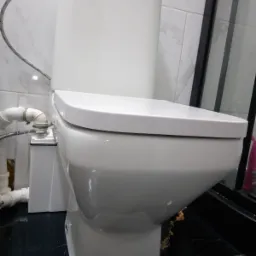 Nowoczesna, biała toaleta kompaktowa z podłączonymi rurami i wężykiem, na tle marmurowej ściany i czarnej podłogi z połyskiem.