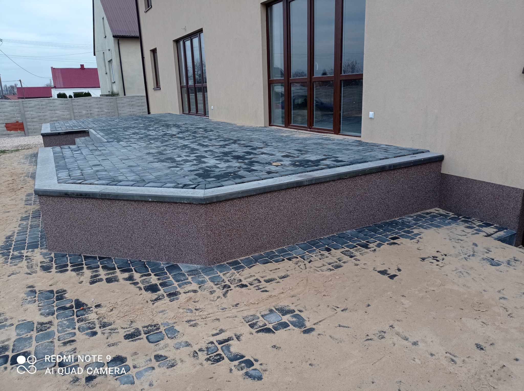 Wykończony taras z kostki brukowej w odcieniach szarości, z widocznymi śladami piasku budowlanego, przylegający do elewacji budynku z dużymi oknami.