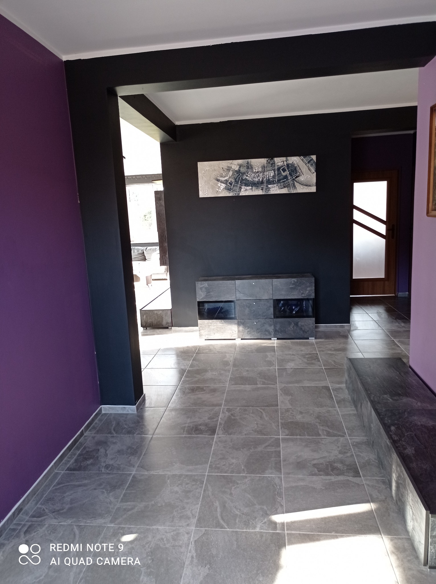 Nowoczesny przedpokój z szarymi płytkami podłogowymi, czarnymi elementami konstrukcyjnymi i fioletowymi ścianami, oświetlony naturalnym światłem wpadającym przez okno.