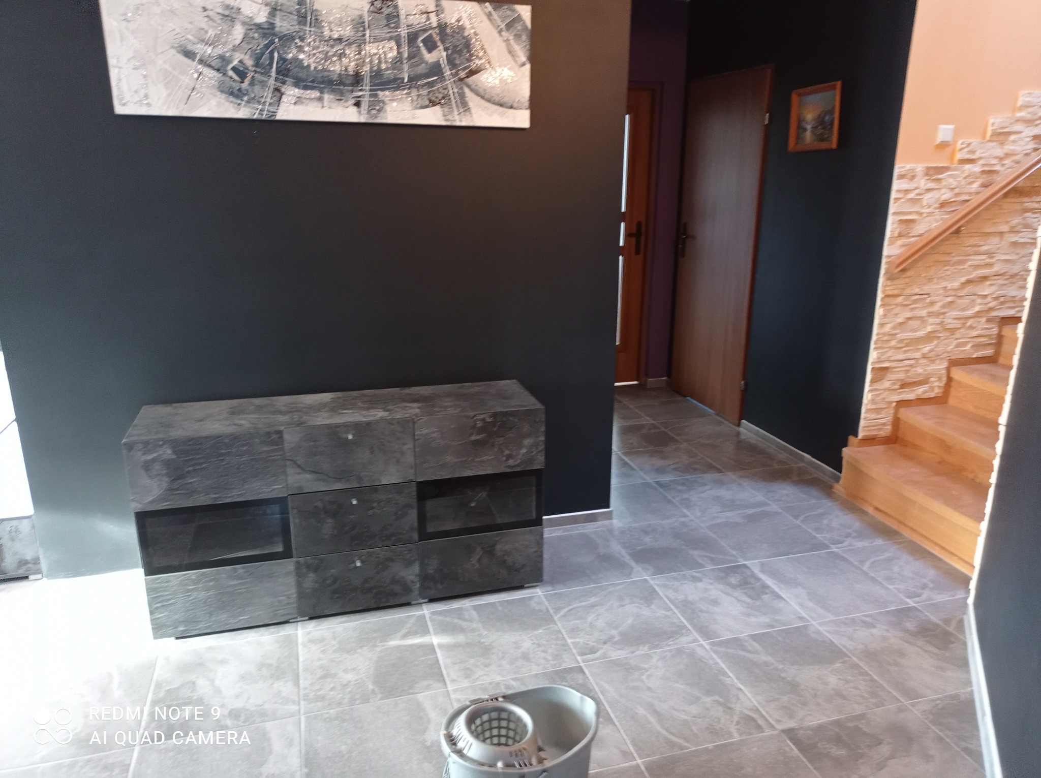 Szare płytki podłogowe ułożone w kwadraty, ciemnoszara ściana z obrazem, komoda w podobnym odcieniu, drewniane schody z kamienną okładziną, widoczne drzwi oraz wiadro z mopem.