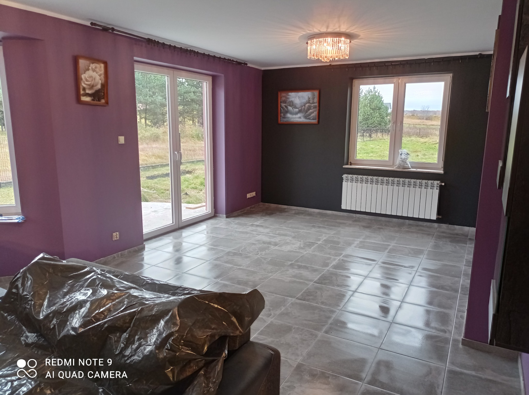Pusty pokój z szarymi płytkami podłogowymi, fioletowymi ścianami, białym grzejnikiem i widokiem na zewnątrz przez okna. Meble przykryte folią ochronną.