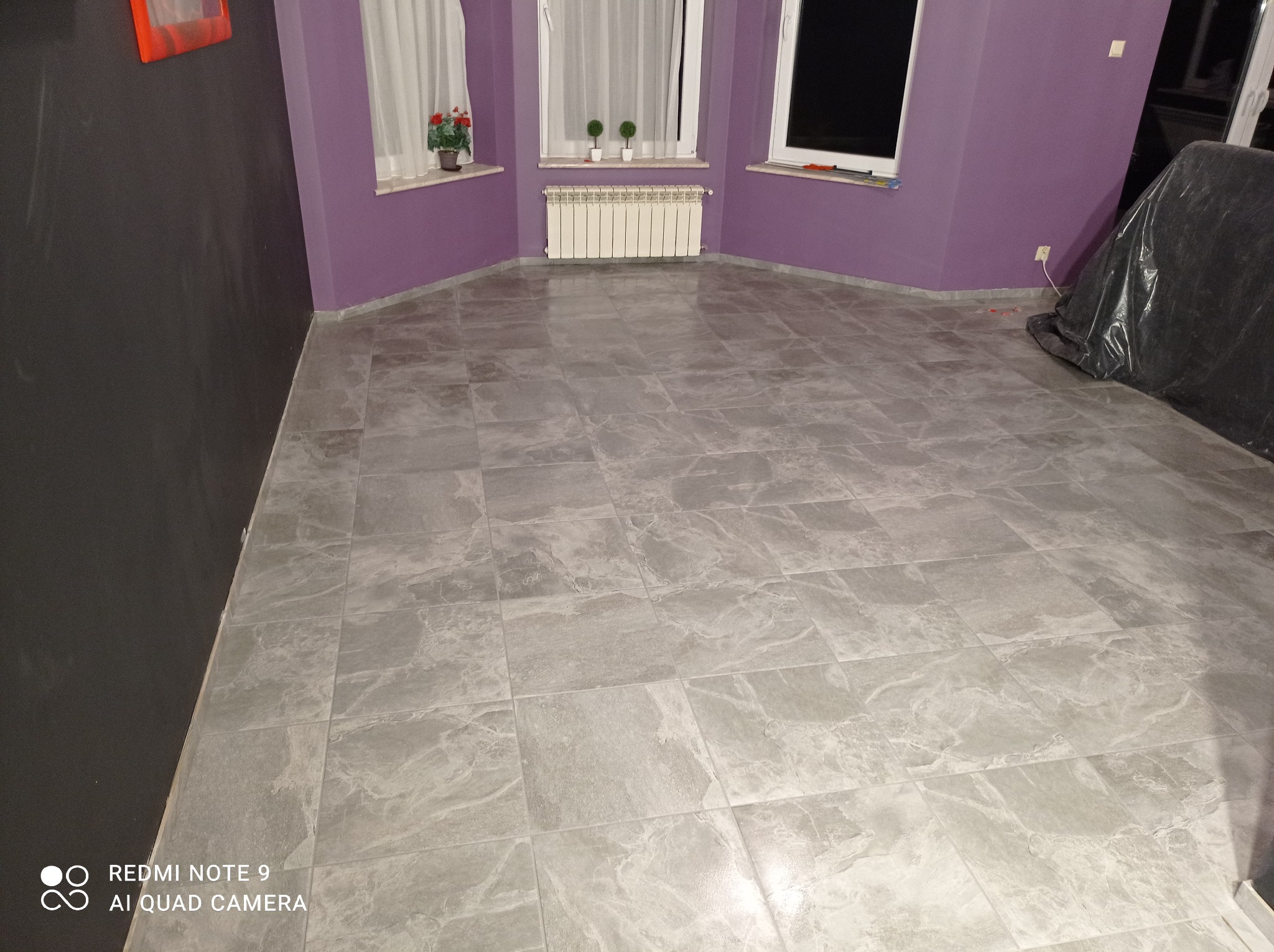 Wykończona podłoga z szarych, kwadratowych płytek imitujących kamień, fioletowe ściany, biały grzejnik pod oknem z białymi firankami, fragment czarnej ściany po lewej, przykryty folią mebel po prawej.