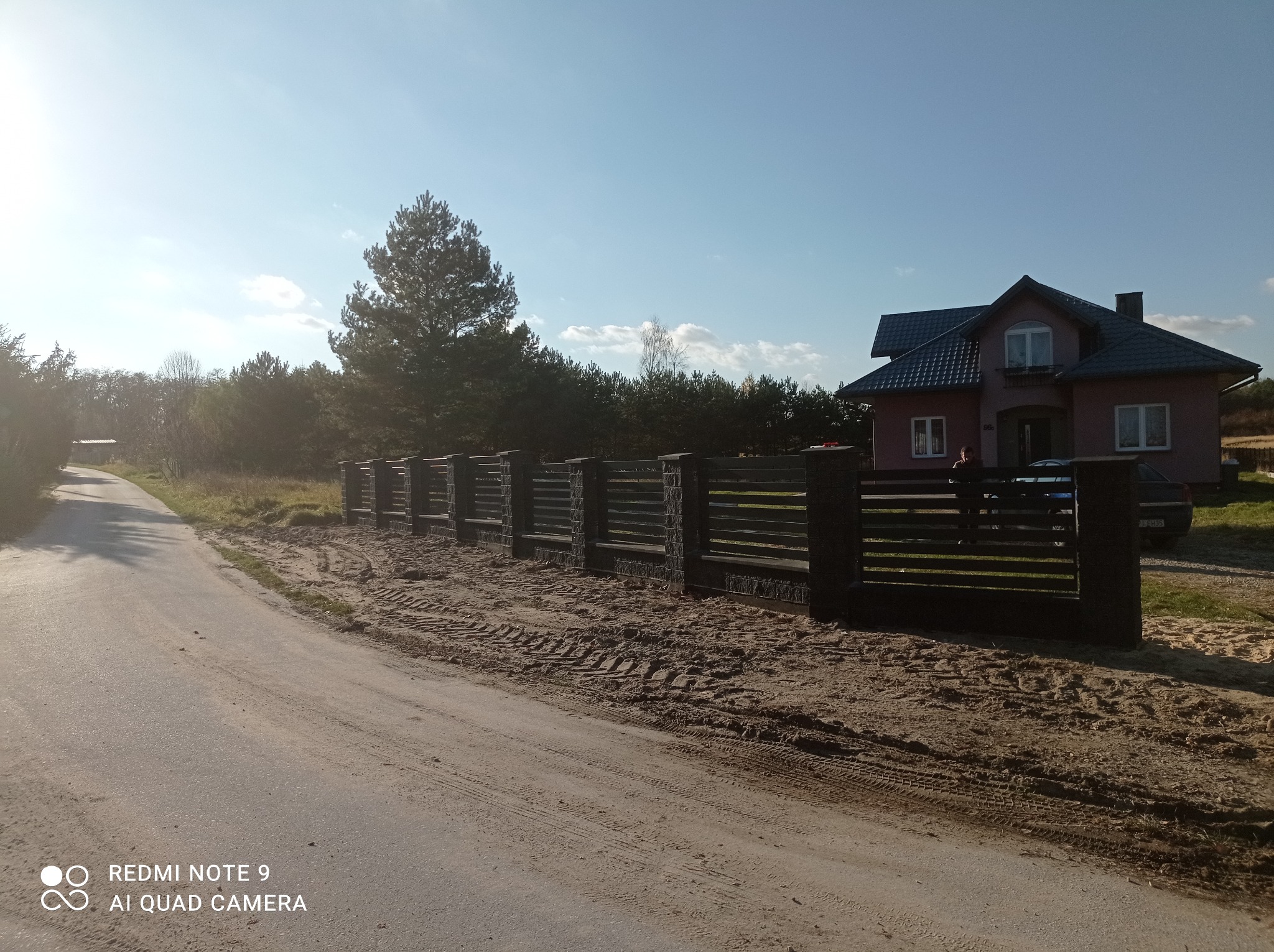 Nowoczesne ogrodzenie panelowe z elementami imitującymi kamień, widoczne świeże ślady opon, dom jednorodzinny w tle, słoneczny dzień.