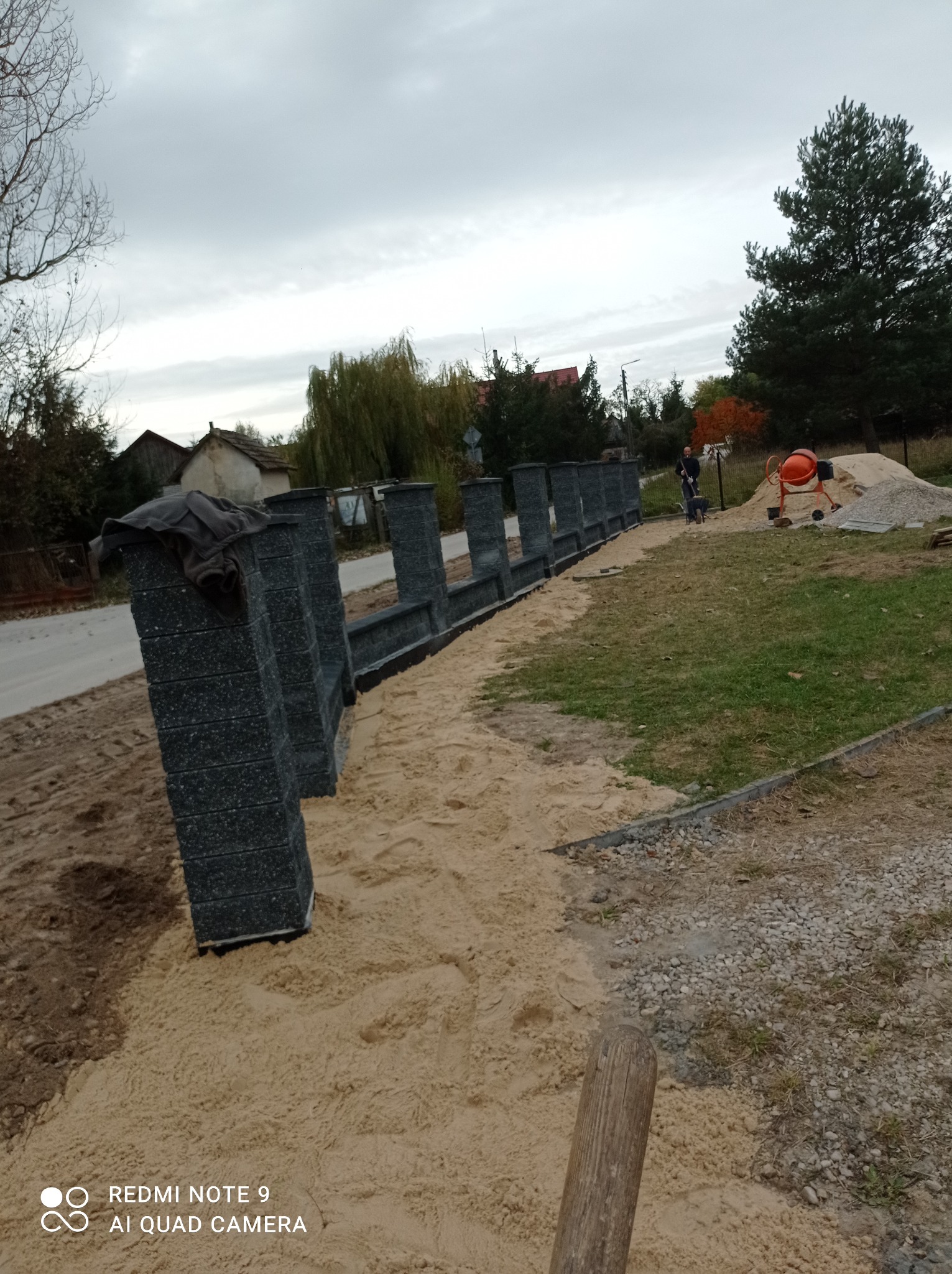 Wznoszony mur z ciemnych, betonowych bloczków na podsypce piaskowej, widoczna betoniarka i pracownik z taczką w tle, pochmurne niebo.