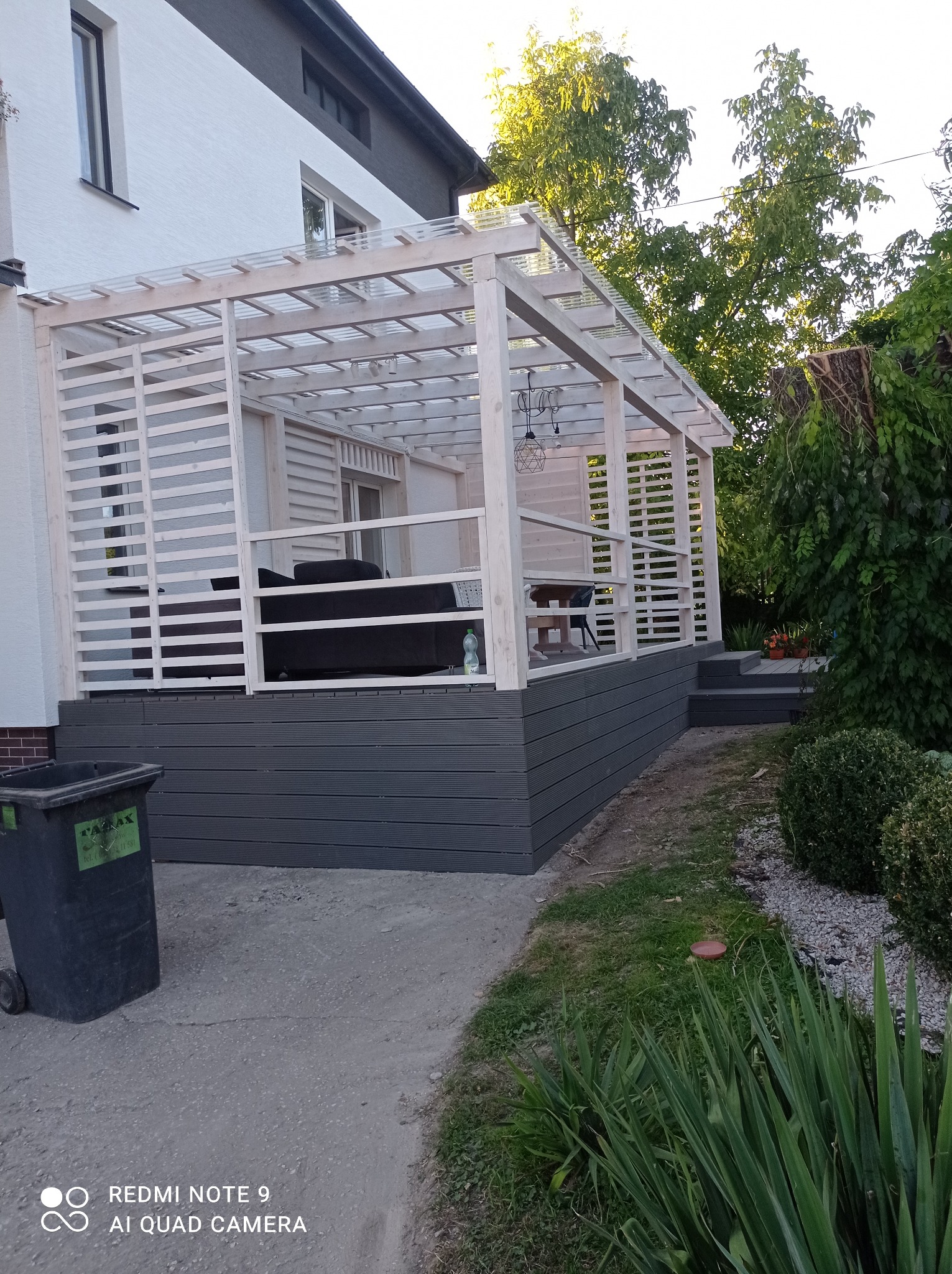 Drewniana pergola metalowa z przezroczystym zadaszeniem przy białym domu w Pińczowie, widoczny fragment tarasu z meblami ogrodowymi i schodami.