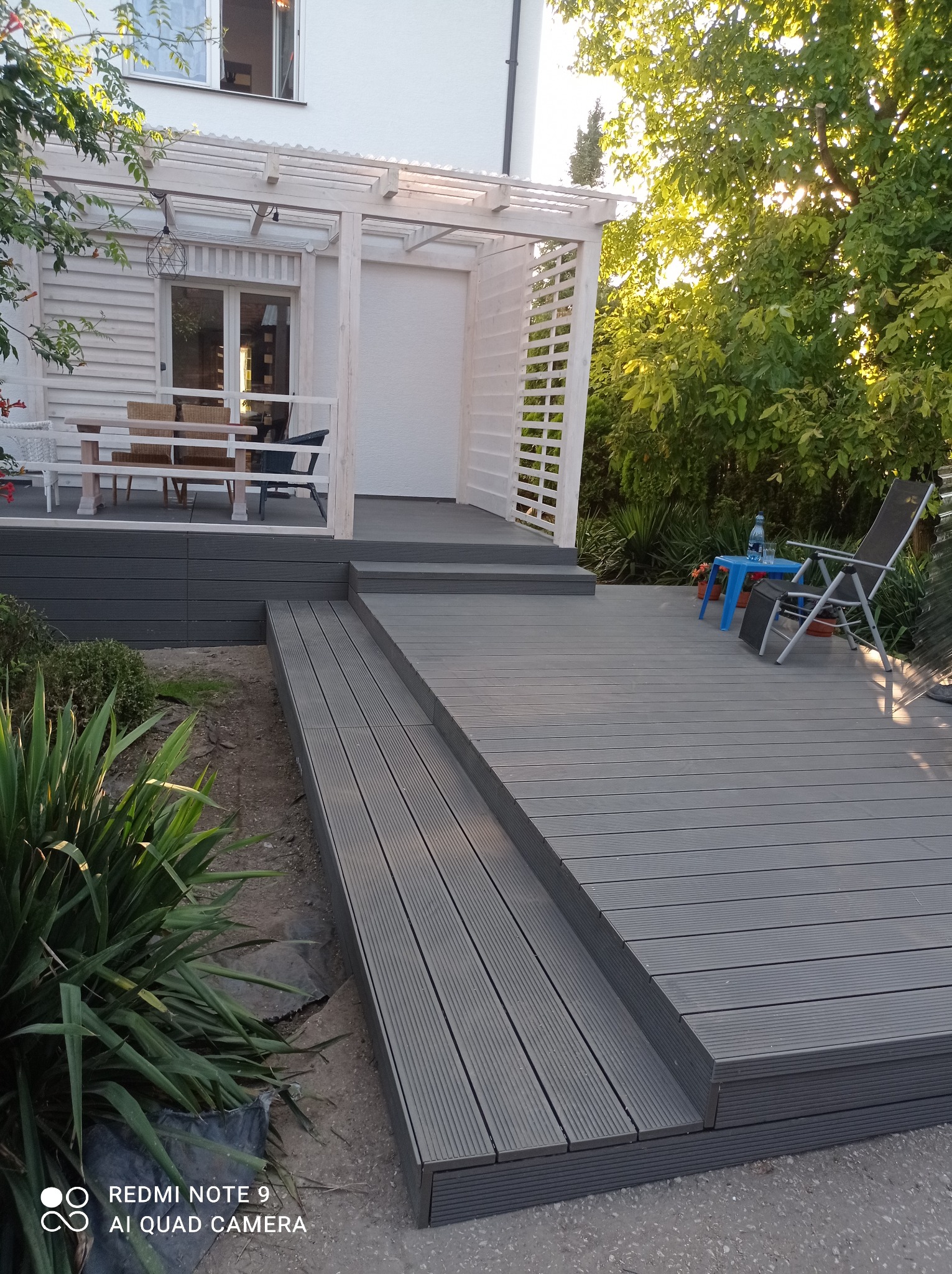 Szara kompozytowa deska tarasowa z niskim stopniem i schodami, prowadząca do białej pergoli z meblami ogrodowymi w tle, otoczona zielenią.