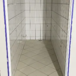 Wnętrze kabiny prysznicowej z białymi płytkami na ścianach i beżowymi na podłodze ułożonymi w karo, widoczne otwory na armaturę i odpływ w podłodze.