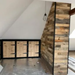 Adaptacja poddasza: zabudowa skosów z drewnianych desek, szafki z jasnego drewna z czarnymi okuciami, surowa podłoga w trakcie prac wykończeniowych.