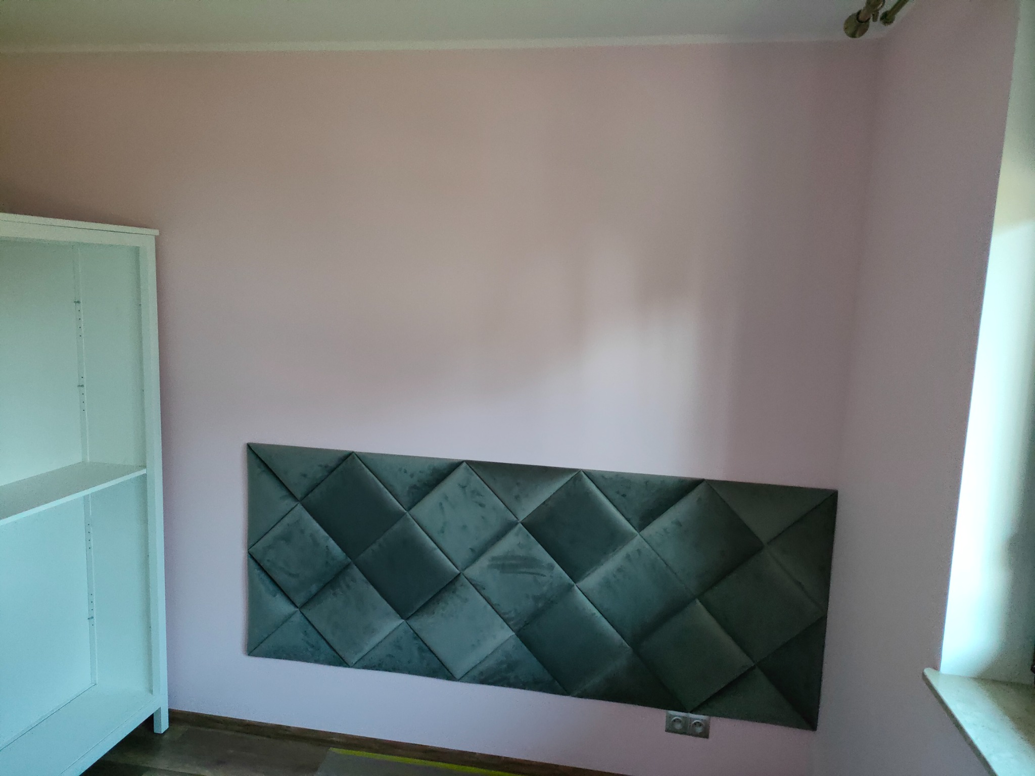 Panel tapicerowany w romby, zamontowany na różowej ścianie w pokoju, obok białego regału.