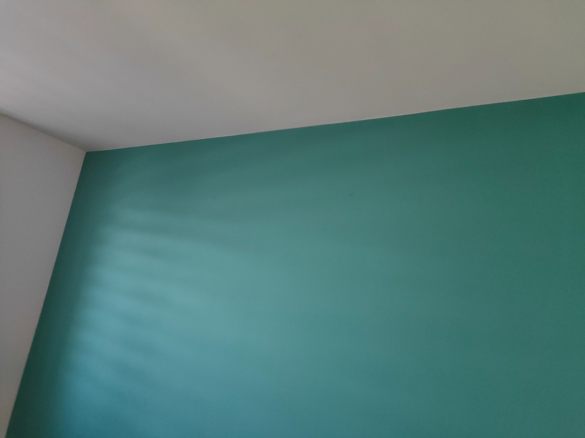 Świeżo pomalowany narożnik pokoju z turkusową ścianą i białą ścianą przylegającą, widoczne delikatne smugi światła na ścianie.