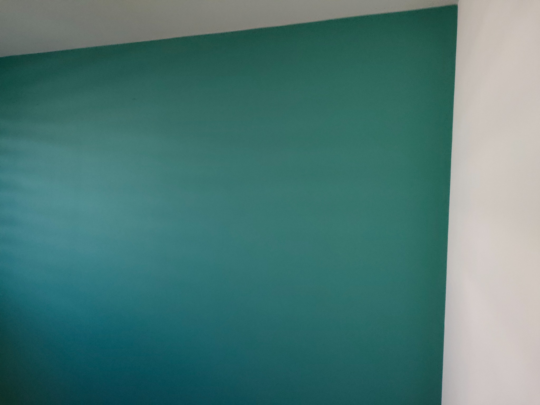 Ściana pomalowana na jednolity, turkusowy kolor, kontrastująca z białą ścianą narożną, widoczne delikatne refleksy światła.