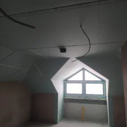 Poddasze w trakcie adaptacji: widoczne płyty kartonowo-gipsowe na skośnym suficie, okno dachowe z nieotynkowanym murem pod spodem oraz wiszące przewody elektryczne.