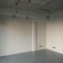 Grzegorz Kowalik - Wnętrze po szpachlowaniu ścian, z widocznym surowym betonowym sufitem z industrialnym oświetleniem i szarą podłogą z płytek.