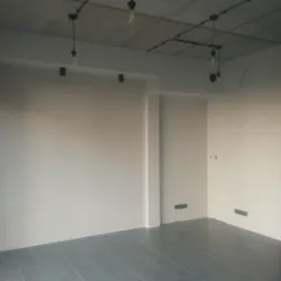 Wnętrze po szpachlowaniu ścian, z widocznym surowym betonowym sufitem z industrialnym oświetleniem i szarą podłogą z płytek.