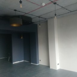 Grzegorz Kowalik - Wnętrze w trakcie prac wykończeniowych: ściany pomalowane na biało i granatowo, widoczne lampy wiszące z żarówkami na kablach, surowy betonowy sufit z instalacją elektryczną na wierzchu, ciemna...