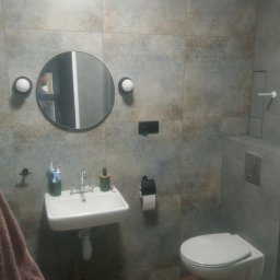 Grzegorz Kowalik - Nowoczesna łazienka z okrągłym lustrem podświetlonym kulistymi lampami, wiszącą umywalką i toaletą w zabudowie, wykończona płytkami w odcieniach szarości i brązu oraz podłogą imitującą drewno.