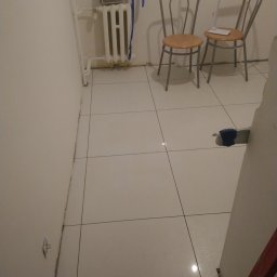 Grzegorz Kowalik - Świeżo ułożone, białe, błyszczące płytki podłogowe w trakcie remontu, widoczne narzędzia i poziomica przy ścianie, dwa krzesła i lampa w tle, zabezpieczenia okna folią malarską.